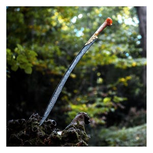 May include: A curved, silver-colored sword with a gold-colored hilt and a red handle. The sword is resting on a tree stump in a forest setting.