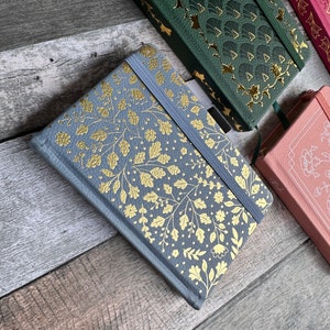 Peut inclure: Trois carnets différents avec des motifs dorés en relief sur les couvertures. Les carnets sont gris, vert et rose. Le carnet gris a un motif floral, le carnet vert a un motif géométrique et le carnet rose a un motif floral.