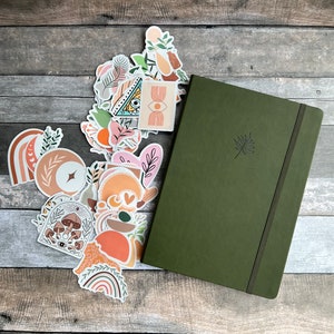 May include: A green hardback journal with a leaf design on the cover. The journal is surrounded by a variety of colourful stickers with abstract designs, including rainbows, moons, and mushrooms.