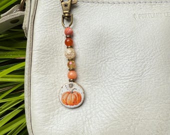 May include: A fall-themed keychain charm with a pumpkin design. The charm features a round, white pendant with an orange pumpkin illustration. It is strung with various beads in shades of orange, peach, and gold, and attached to a bronze-colored clasp.