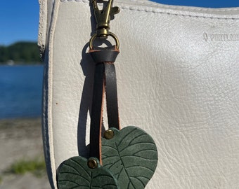 Puede incluir: Un dije de hoja de cuero verde oscuro con vetas detalladas, sujeto a un cierre de color bronce y una correa de cuero marrón oscuro. El dije cuelga de un bolso blanco con un cierre de color bronce.