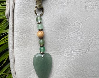 May include: A green heart-shaped charm with a beaded chain attached to a gold-toned metal clasp. The charm is hanging from a white leather bag.