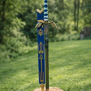 Op de afbeelding: Een gedetailleerd replica zwaard met een blauw en goudkleurig gevest en lemmet, rustend op een houten stronk. Het zwaard heeft een blauwe handgreep met een kruispatroon en een gouden pareerstang met vleugelachtige details. De schede is blauw met gouden accenten.