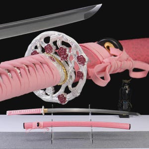 Op de afbeelding: Een Japanse katana-zwaard met een roze handvat en schede. Het zwaard heeft een zilveren beschermkap met rode bloemaccenten. Het lemmet is zilverkleurig en het handvat is omwikkeld met roze stof. Een klein zwart samoerai-figuur is ook zichtbaar.