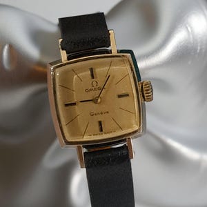 Pode incluir: Um relógio Omega Genève vintage com mostrador quadrado dourado e pulseira de couro preta. O mostrador exibe o logotipo da Omega e a palavra "Genève". O relógio tem ponteiros e marcadores de hora dourados. Um relógio clássico.