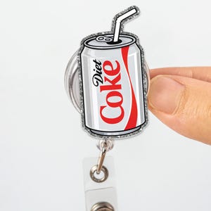 May include: A Diet Coke can-shaped badge reel with a silver glitter border. The can is white with the Diet Coke logo in red. A white straw extends from the top of the can. The reel has a clear plastic strap and a metal clip.
