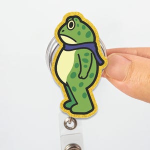 May include: A green frog badge reel with a gold glitter border. The frog has a blue scarf and a sad expression. The badge reel is white and silver.