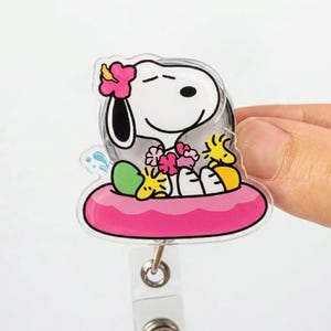 Puede incluir: Un portatarjetas retráctil transparente de Snoopy con Snoopy en un flotador rosa con flores. El portatarjetas tiene un diseño rosa y blanco con una parte trasera de plástico transparente y un clip de metal.