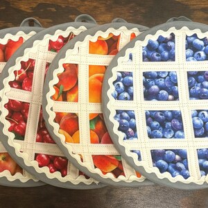 Könnte beinhalten: Vier runde Topflappen mit einem Kuchendesign. Jeder Topflappen hat einen grauen Rand und ein weißes Gittermuster über einer anderen Fruchtfüllung: Äpfel, Kirschen, Pfirsiche und Blaubeeren. Die Topflappen haben eine Schlaufe zum Aufhängen.