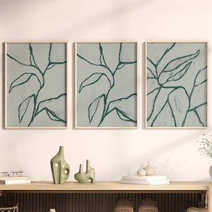 Op de afbeelding: Een lange houten consoletafel met verticale latten en een open plank, voorzien van drie ingelijste wandkunstwerken met donkergroene abstracte bladlijn-tekeningen op een lichtgrijze achtergrond, plus diverse decoratieve vazen en een strooien hoed.