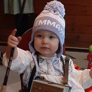 May include: A young child wearing a light blue and white knit hat with the name "EMMA" on it. The child is also wearing a white shirt and holding a dark-colored object. The child is sitting in a chair.
