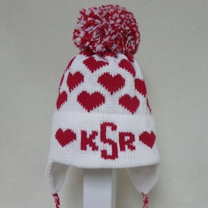 May include: White knit hat with red hearts and a red pom pom. The hat has ear flaps and a long white and red braided cord with a tassel. The hat has the letters "KSR" knitted in red on the front.