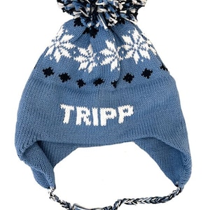 May include: Blue knitted winter hat with a white pom pom and snowflake pattern. The hat has ear flaps and a long tassel. The name "TRIPP" is embroidered on the front of the hat.