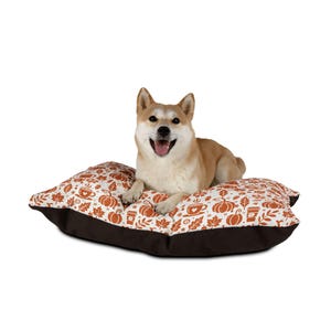 May include: A brown and white dog bed with a fall-themed print of pumpkins, leaves, and coffee cups. A light brown dog is lying on the bed.