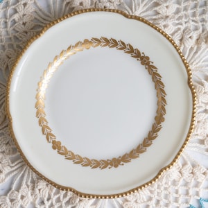 May include: A white porcelain plate with a gold rim and a gold leaf design around the edge.