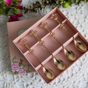 May include: A set of six gold-tone teaspoons with crown-shaped handles, arranged in a pink box with floral accents.