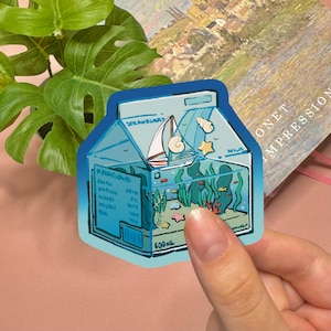 May include: A die-cut sticker shaped like a milk carton with a blue gradient border. The illustration depicts an underwater scene inside the carton featuring a sailboat, seaweed, coral, and seashells. The text reads Strawberry, Milk, and 500ml.