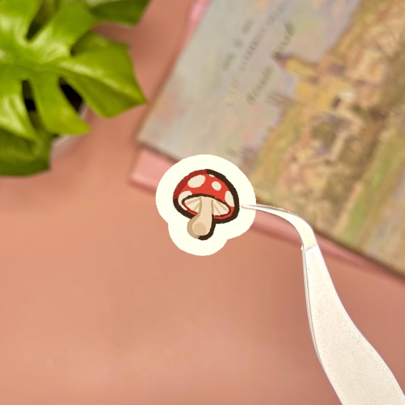 May include: A small, circular sticker featuring a red mushroom with white spots and a tan stem. The sticker is held by a pair of silver metal tweezers, showcasing the detailed illustration of the fungus against a plain, light-colored background.