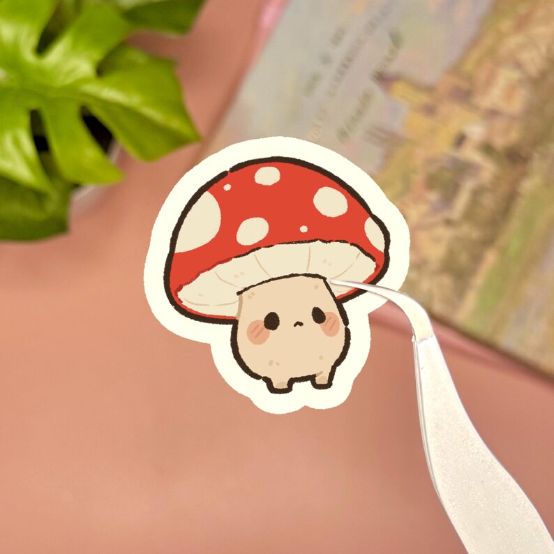 May include: A die-cut sticker featuring a cartoon mushroom character with a red cap adorned with white spots. The mushroom has a beige stem, a small face with black eyes, and pink cheeks. The sticker is held by a pair of silver metal tweezers.