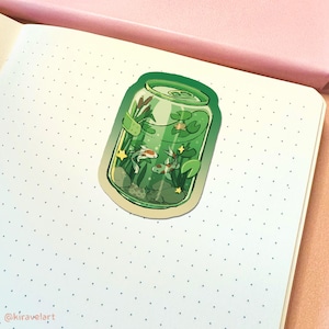 May include: A green sticker in the shape of a soda can, featuring an illustration of a small pond with koi fish, aquatic plants, and lily pads. The sticker has a white border and is placed on a dotted notebook page.