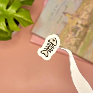 May include: A small, die-cut sticker featuring a stylized illustration of a fish skeleton. The design is rendered in black outlines with a cream-colored fill, and the fish head includes a small 'x' mark for an eye. The sticker is held by silver tweezers.