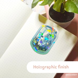 Op de afbeelding: Een holografische sticker van een blikje met een berglandschap erin. Het blikje is blauw en groen met een witte rand. Het berglandschap is een blauw en groen landschap met witte sterren. De sticker heeft een holografische afwerking die in het licht glinstert.