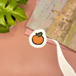 May include: A small, circular sticker featuring a hand-drawn illustration of a bright orange fruit with a dark brown outline and a single green leaf on top. The sticker is held by a pair of silver metal tweezers, showcasing its compact size.