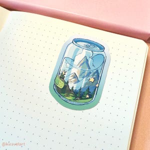 Op de afbeelding: Een sticker van een blikje met een berglandschap erin. Het blikje is blauw en wit met een groen deksel. Het berglandschap is een blauwe berg met een groen bos aan de voet. Er zijn gele sterren verspreid over de sc&egrave;ne.