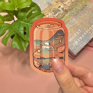 May include: A die-cut sticker shaped like an orange soda can featuring a landscape illustration inside. The design depicts a mountain peak, clouds, and stars in a sunset color palette. The sticker has a smooth, rounded edge and a glossy finish.