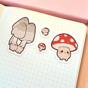 May include: A set of four die-cut stickers featuring anthropomorphic mushrooms. One large cluster of beige mushrooms, two tiny individual mushrooms, and one large red-capped mushroom with white spots, all with simple faces and pink cheeks.