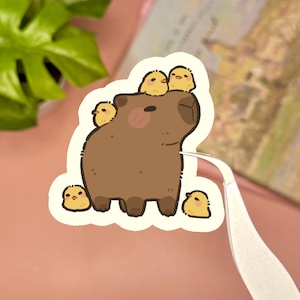 May include: A die-cut sticker featuring a brown capybara with four small yellow chicks. Two chicks are perched on the capybara's head, while one sits on its back and another stands at its feet. The illustration has a white border and a matte finish.
