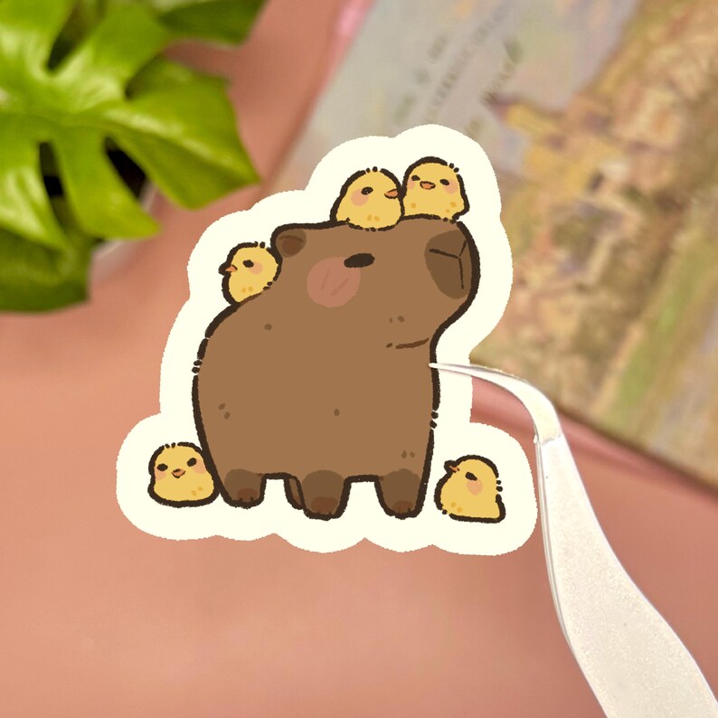 May include: A die-cut sticker featuring a brown capybara with four small yellow chicks. Two chicks are perched on the capybara's head, while one sits on its back and another stands at its feet. The illustration has a white border and a matte finish.