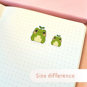 May include: Two green frog stickers with a small plant on their heads. The stickers are on a white dotted background. The text "Size difference" is written on a pink rectangle at the bottom of the image.
