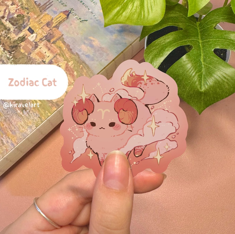 May include: A pink cat with ram horns and a fluffy tail, surrounded by sparkling stars. The cat is a zodiac sign illustration, with the text "Zodiac Cat" and "@kiravelart" below.