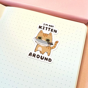 May include: A die-cut sticker featuring an orange tabby cat with large eyes holding a silver kitchen knife in its mouth. The text above the cat reads I'M NOT and the text below reads KITTEN AROUND in dark brown capital letters on a white background.