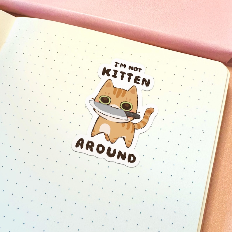 May include: A die-cut sticker featuring an orange tabby cat with large eyes holding a silver kitchen knife in its mouth. The text above the cat reads I'M NOT and the text below reads KITTEN AROUND in dark brown capital letters on a white background.