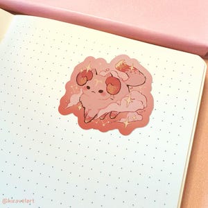 May include: A pink and orange sticker featuring a cartoon cat with ram horns, surrounded by clouds and stars. The cat is wearing a pink and orange outfit and has a cute expression on its face.