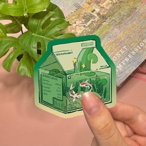 May include: A die-cut sticker shaped like a milk carton filled with water, green aquatic plants, lily pads, and two koi fish. The carton features the text 'STRAWBERRY', 'MILK', and '500ml' printed in a dark green font against a light green background.