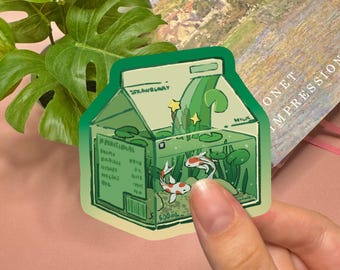 May include: A die-cut sticker shaped like a milk carton filled with water, green aquatic plants, lily pads, and two koi fish. The carton features the text 'STRAWBERRY', 'MILK', and '500ml' printed in a dark green font against a light green background.