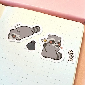 May include: A set of four die-cut stickers featuring cartoon raccoons. One raccoon is sleeping with zzz text, another holds a white daisy with a small heart, accompanied by a black trash bag sticker and a white fish bone skeleton sticker.