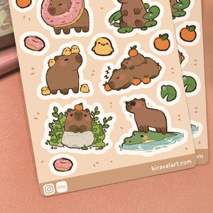 May include: A sticker sheet featuring brown capybaras in various scenes, including one with a pink donut, one with yellow chicks, one sleeping with oranges, one on a rock with greenery, and one standing on a green alligator. The text reads kiravelart.com.