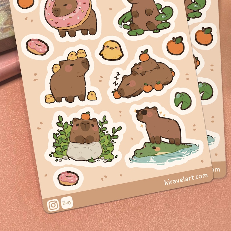 May include: A sticker sheet featuring brown capybaras in various scenes, including one with a pink donut, one with yellow chicks, one sleeping with oranges, one on a rock with greenery, and one standing on a green alligator. The text reads kiravelart.com.