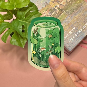 May include: A die-cut sticker featuring a green aluminum soda can filled with water, aquatic plants, cattails, lily pads, and two koi fish. The illustration includes small yellow star accents and a light green gradient background within the can.