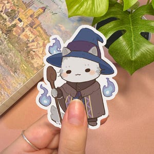 May include: A die-cut sticker featuring a cartoon cat dressed as a wizard in a dark blue pointed hat and brown cloak. The cat holds a wooden staff, surrounded by small, light blue floating flames against a white border outline.