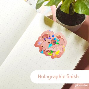 May include: Holographic sticker featuring a pink cartoon character with horns and a cloud background. The sticker has a rainbow iridescent finish. The text "Holographic finish" is printed below the sticker. The text "@kiravelart" is printed in the bottom right corner.