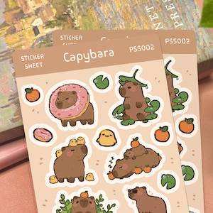 May include: A beige sticker sheet labeled Capybara PSS002 featuring various illustrations of brown capybaras. The designs include a capybara with a pink donut, a capybara with lily pads, a capybara with small yellow chicks, and oranges.