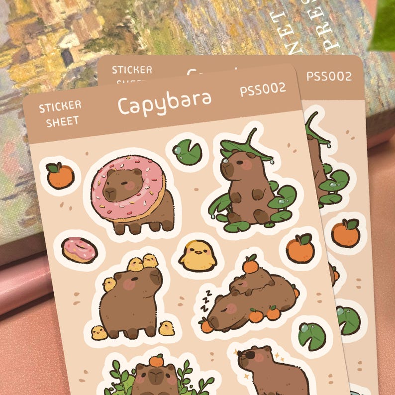 May include: A beige sticker sheet labeled Capybara PSS002 featuring various illustrations of brown capybaras. The designs include a capybara with a pink donut, a capybara with lily pads, a capybara with small yellow chicks, and oranges.