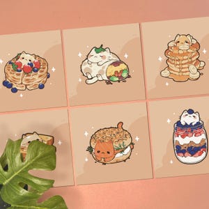 May include: Six square art prints featuring cartoon cats integrated with food items: a waffle with berries, a burrito, a stack of banana pancakes, a sandwich, a bagel with salmon, and a fruit parfait in a glass jar, all on a light tan background.