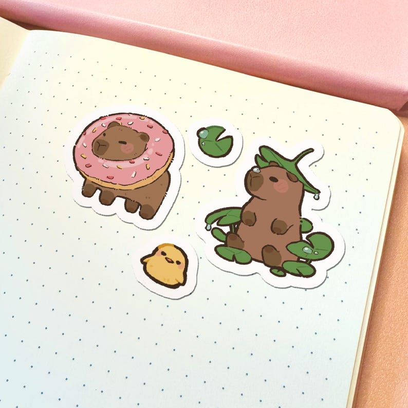 May include: A set of die-cut stickers featuring a brown capybara wearing a pink frosted donut, a small yellow chick, a single green lily pad, and a brown capybara sitting among several green lily pads with one resting on its head.