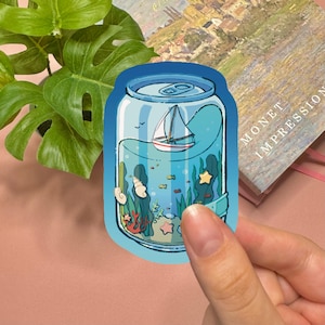 May include: A die-cut sticker shaped like a soda can featuring an underwater scene inside. The illustration shows a small white sailboat floating on the surface, with green seaweed, orange coral, seashells, and a yellow starfish at the bottom.